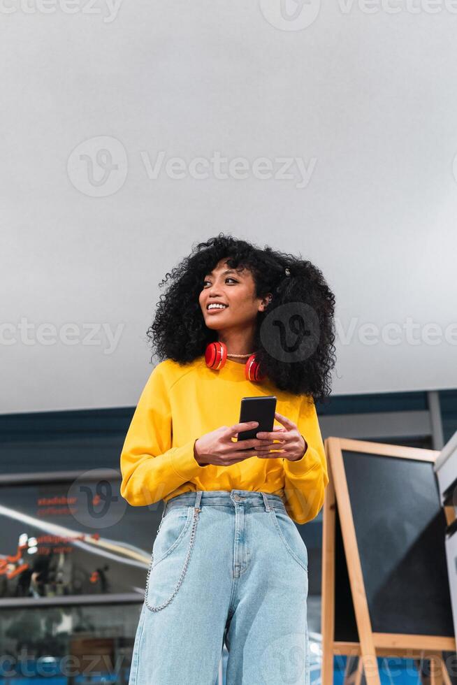 Cheerful stylish ethnic lady standing and using smartphone in contemporary building photo