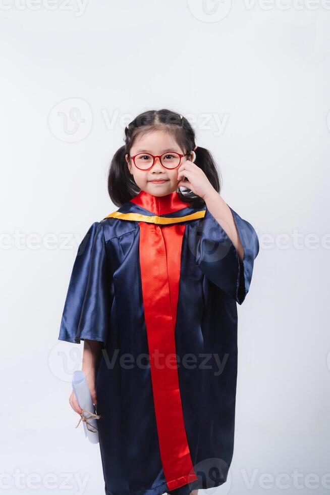 Joyful Asian young girl in graduation gown with red glasses holding a diploma and making a phone call against a plain background photo