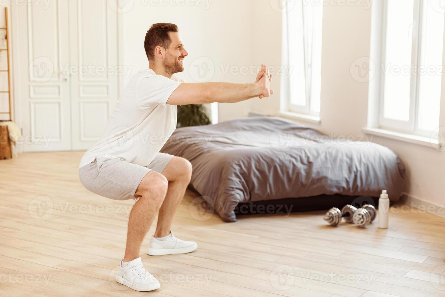Man Doing Two-Legged Squat Exercise Training At Home. Sporty Lifestyle, Male Fitness Workout Routine Concept. Guy Exercising In Bedroom In The Morning. Empty Space For Text photo