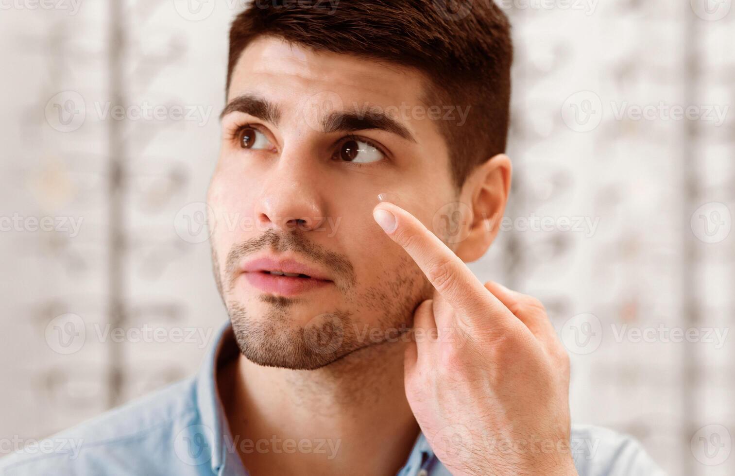 A man with brown hair and a beard is inserting a contact lens in his eye. He is in an eye care shop with a wall of glasses and frames behind him. photo