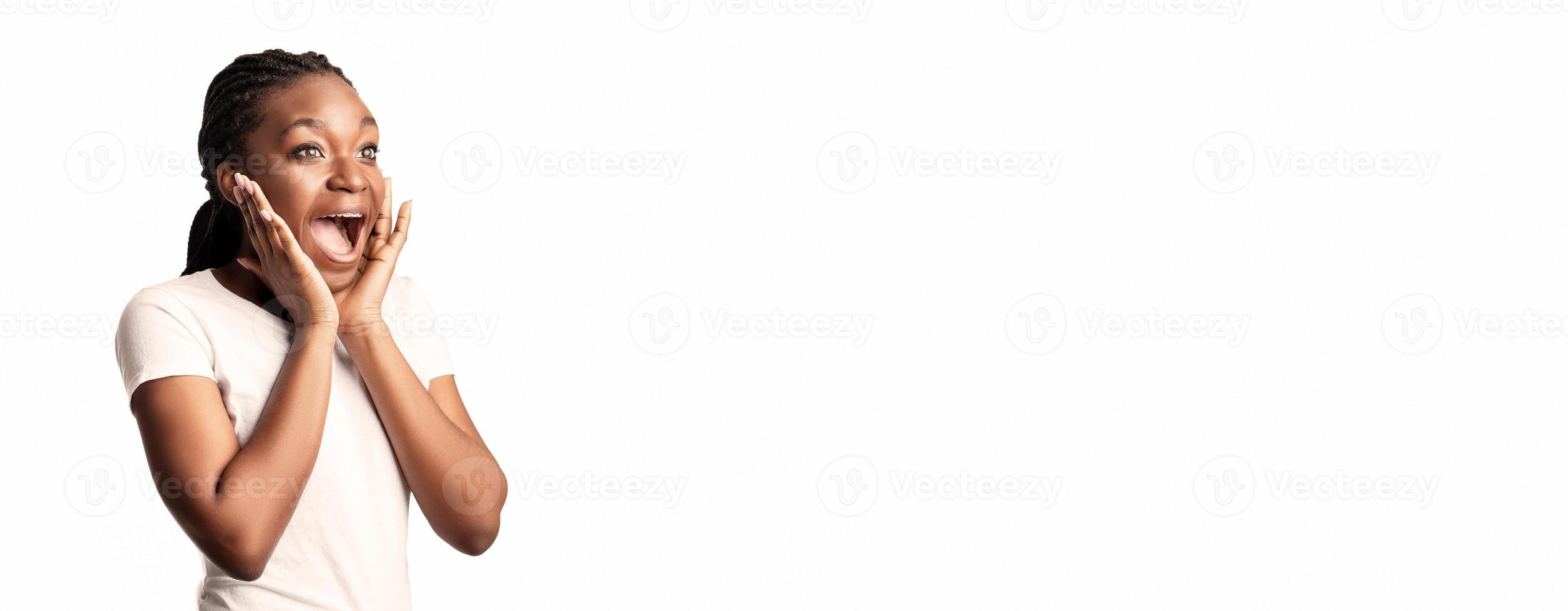 A young lady is exhibiting excitement and surprise, with her hands on her cheeks and a big smile. The bright backdrop emphasizes her joyful expression and playful demeanor. photo