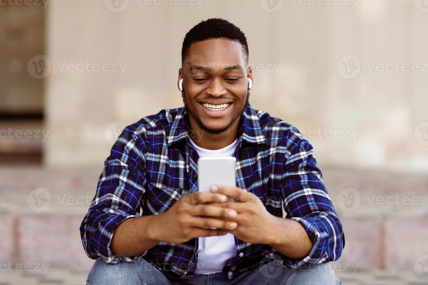 Enjoying Free Time. Happy african hipster wearing headphones and holding cell phone, listening to song in the street photo