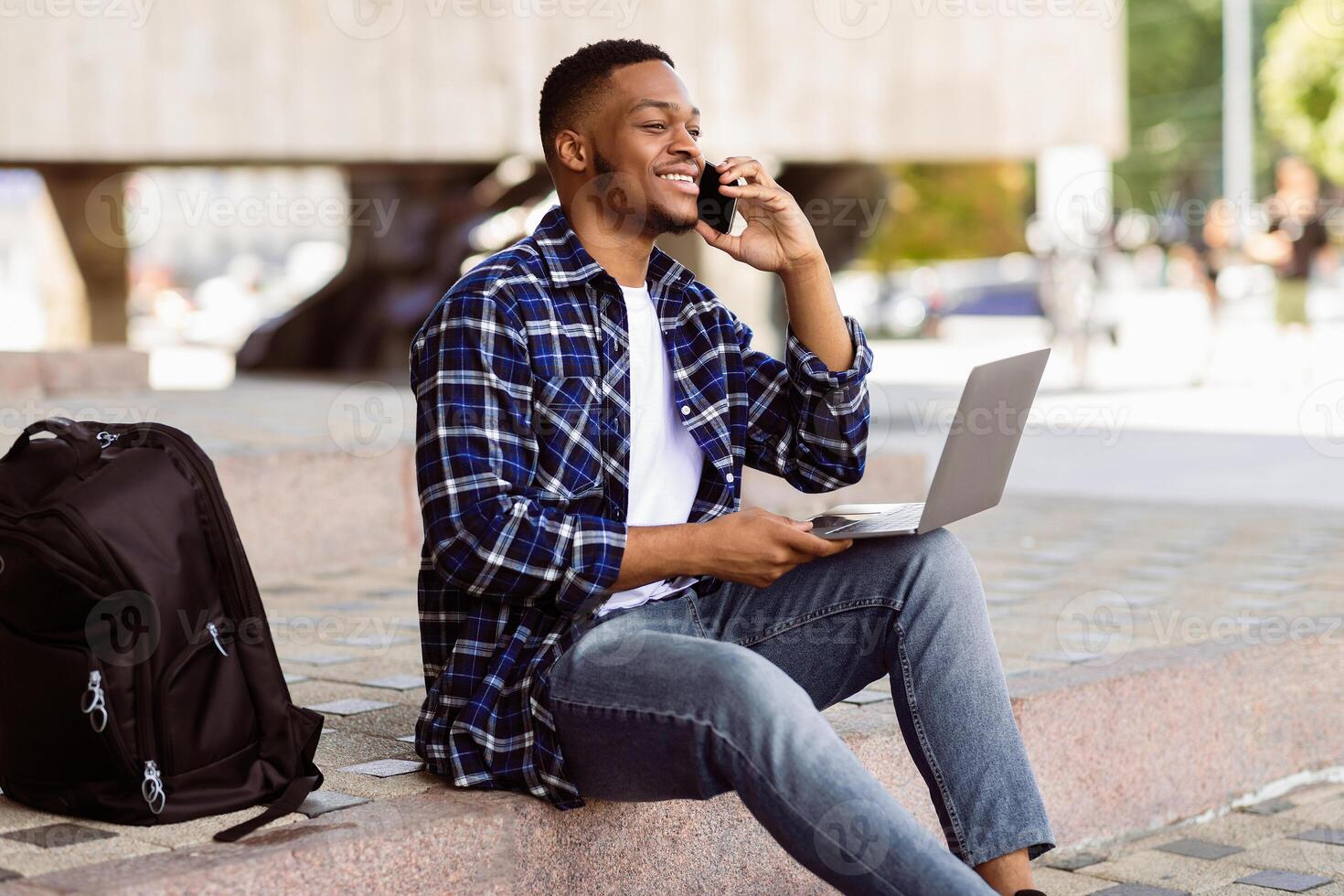 Good Business Talk. Handsome black man talking on smart phone and using laptop while working in the street photo