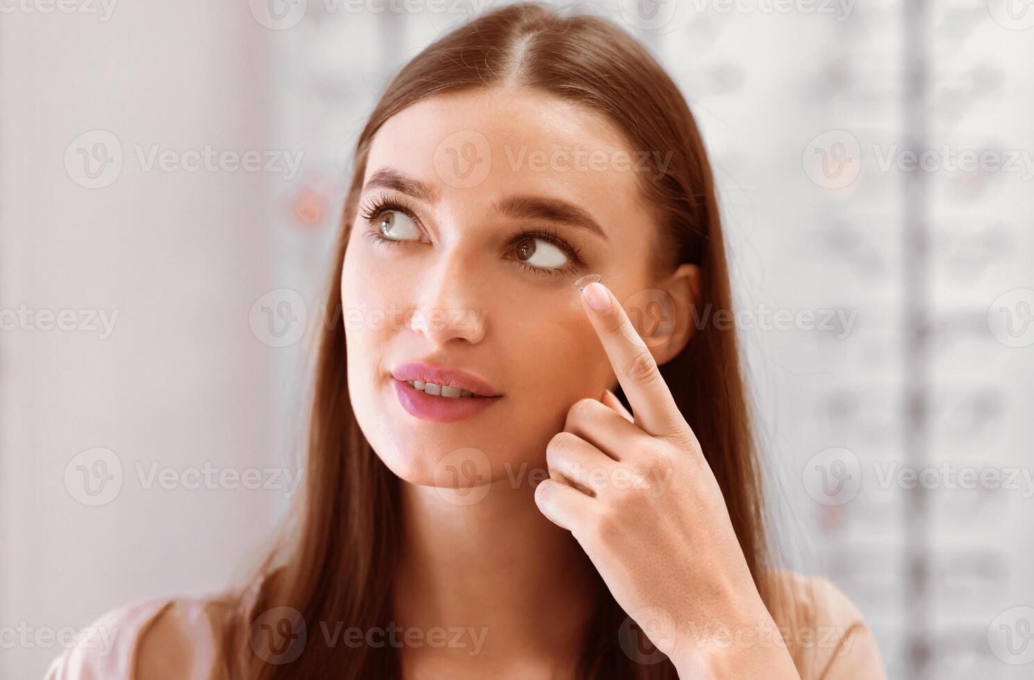 A woman with long, brown hair is inserting a contact lens into her right eye. Her left eye is looking towards the right side of the frame. She is smiling slightly and wearing braces. photo
