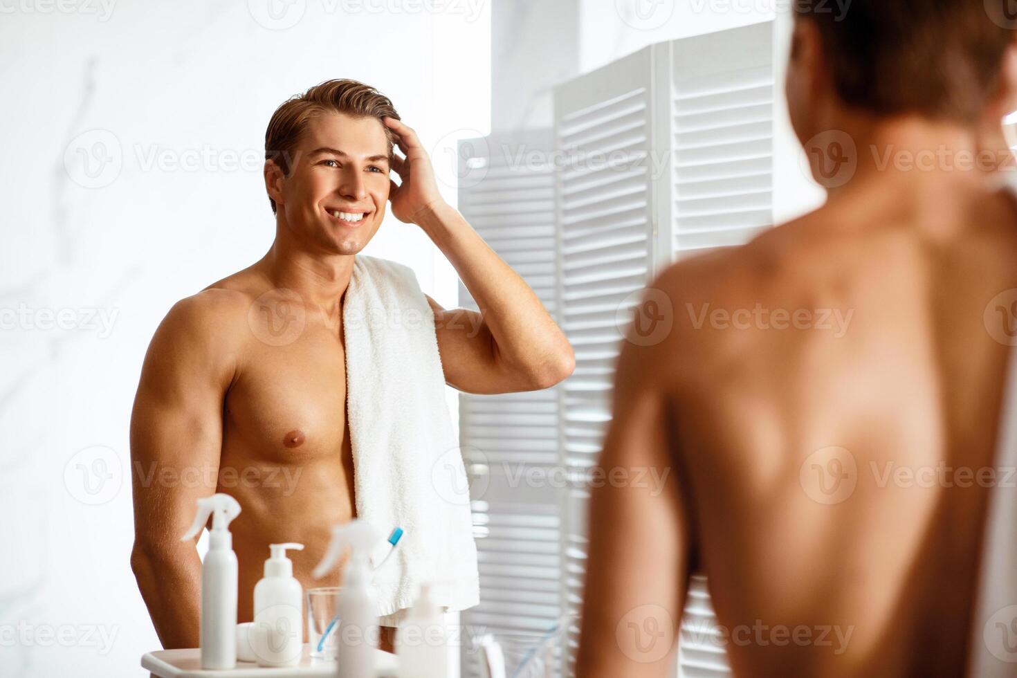 Morning Routine. Handsome young man touching his hair with hand and smiling while standing in front of the mirror photo