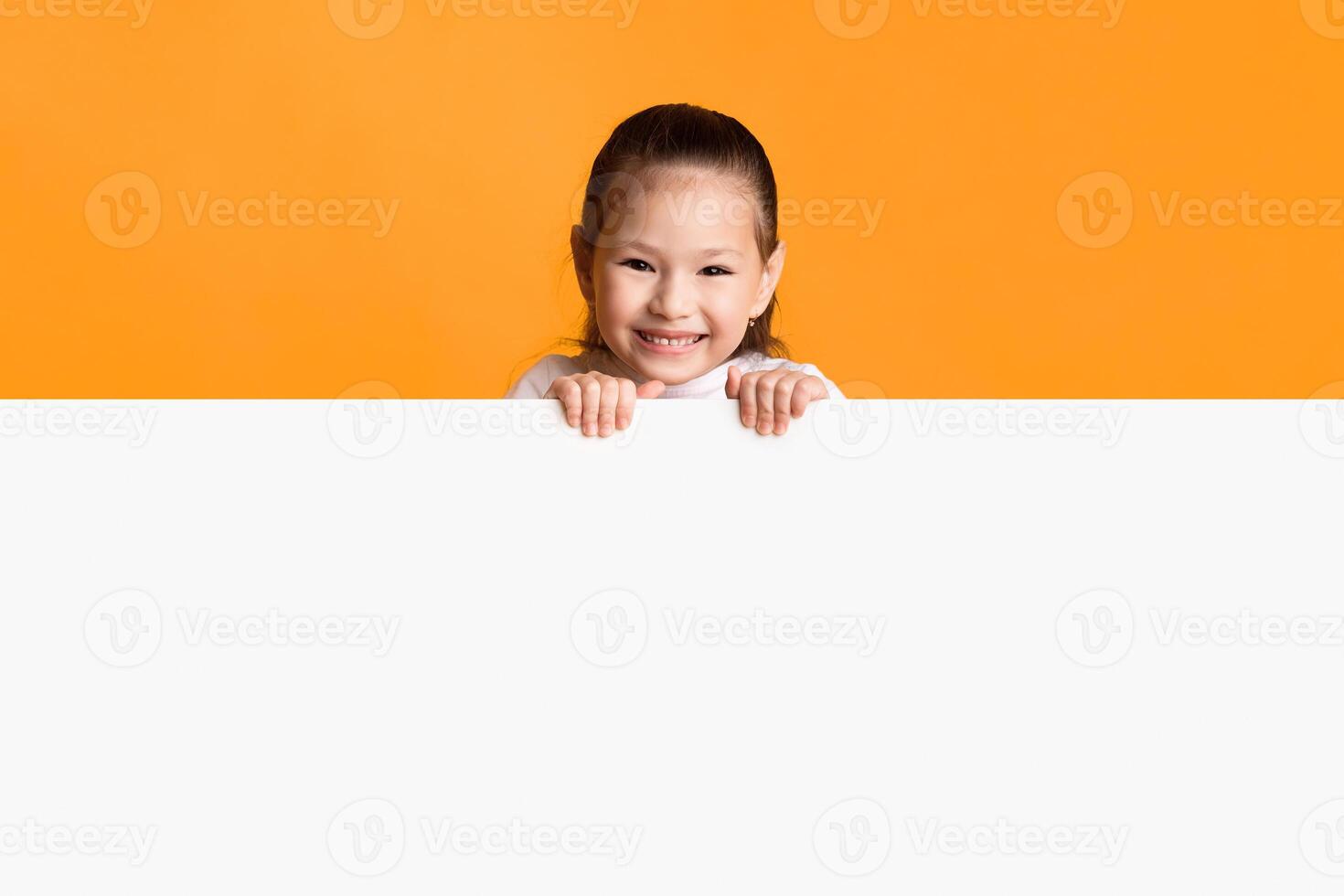 Cute asian kid looking out white advert signboard with empty space, yellow studio wall photo