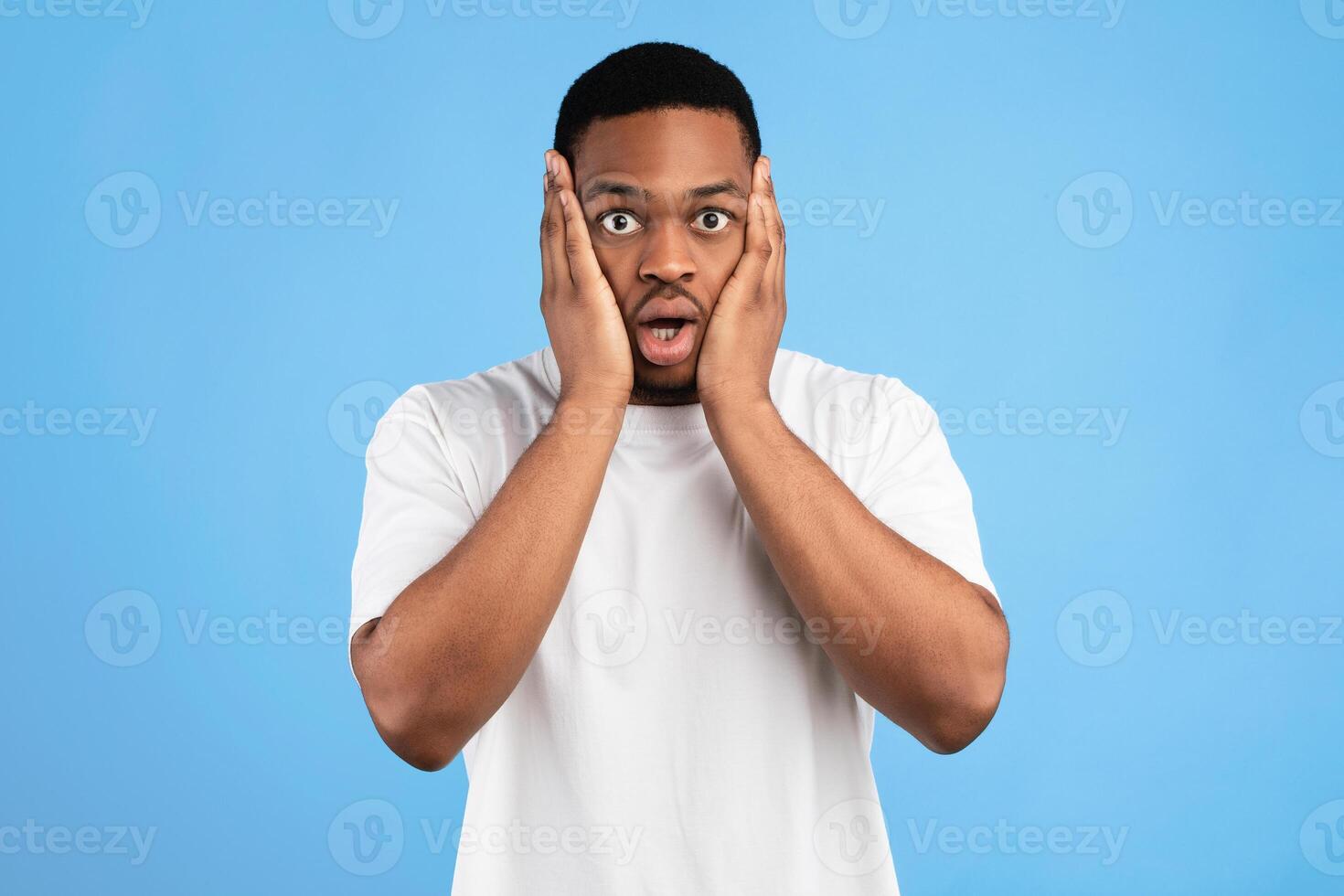 Overwhelmed Black Man Cupping Face In Hands Looking At Camera In Shock Standing On Blue Background. Studio Shot photo