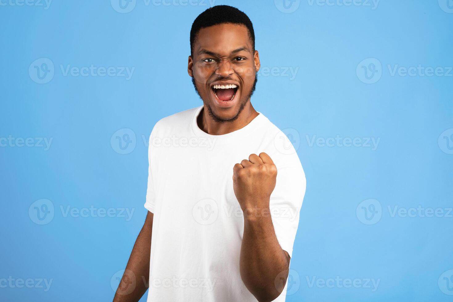Joy Of Victory. African Man Shaking Fist Gesturing Yes Celebrating Success Over Blue Background. Studio Shot photo