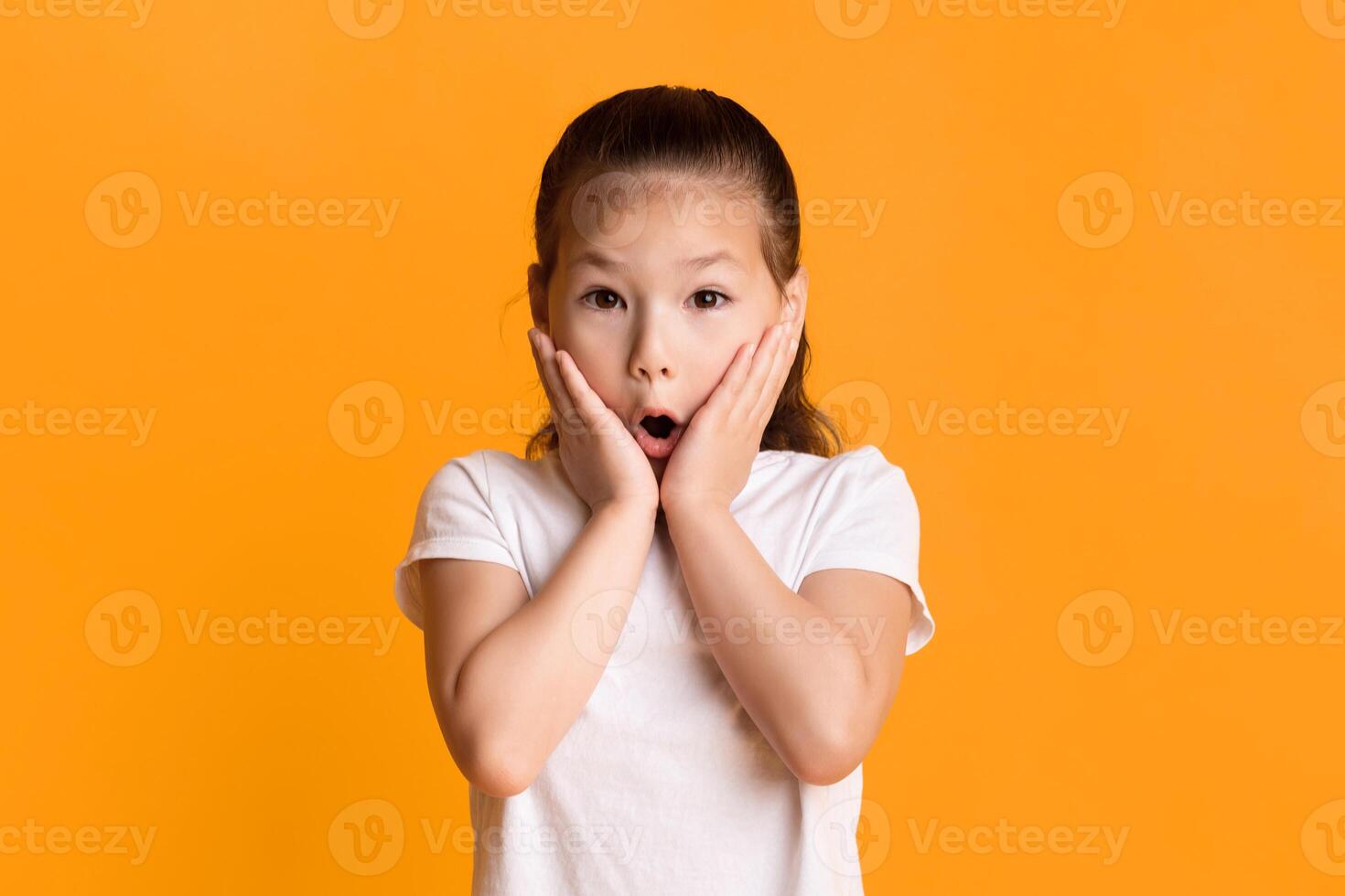 Wow. Portrait of surprised and shocked asian girl touching cheeks with open mouth in amazement isolated over yellow studio background. Child saying omg or oops, no way, looking at camera photo