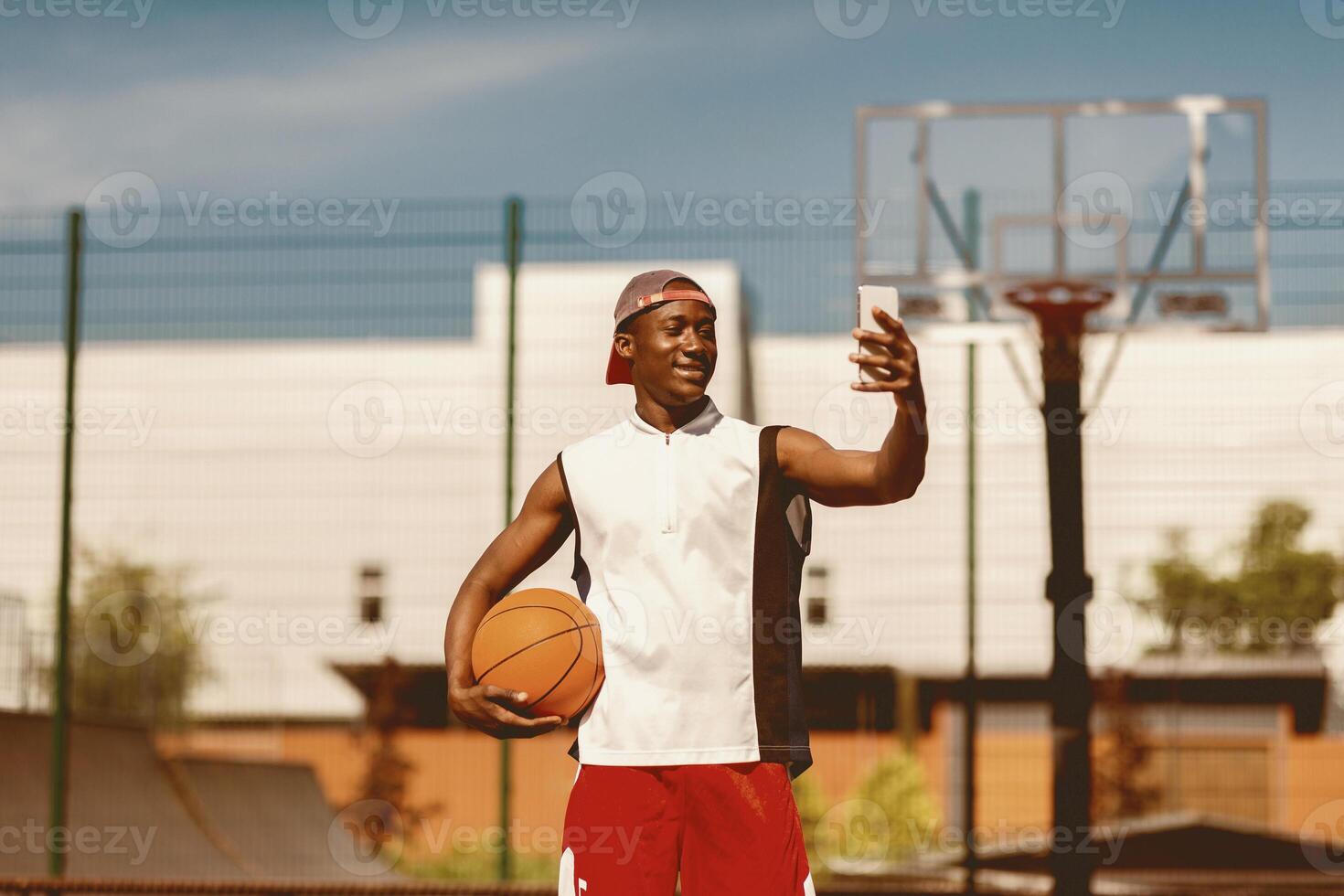 Portrait of black basketball player holding ball, taking selfie on smartphone at outdoor sports arena. Millennial African American sportsman making photo of himself outside