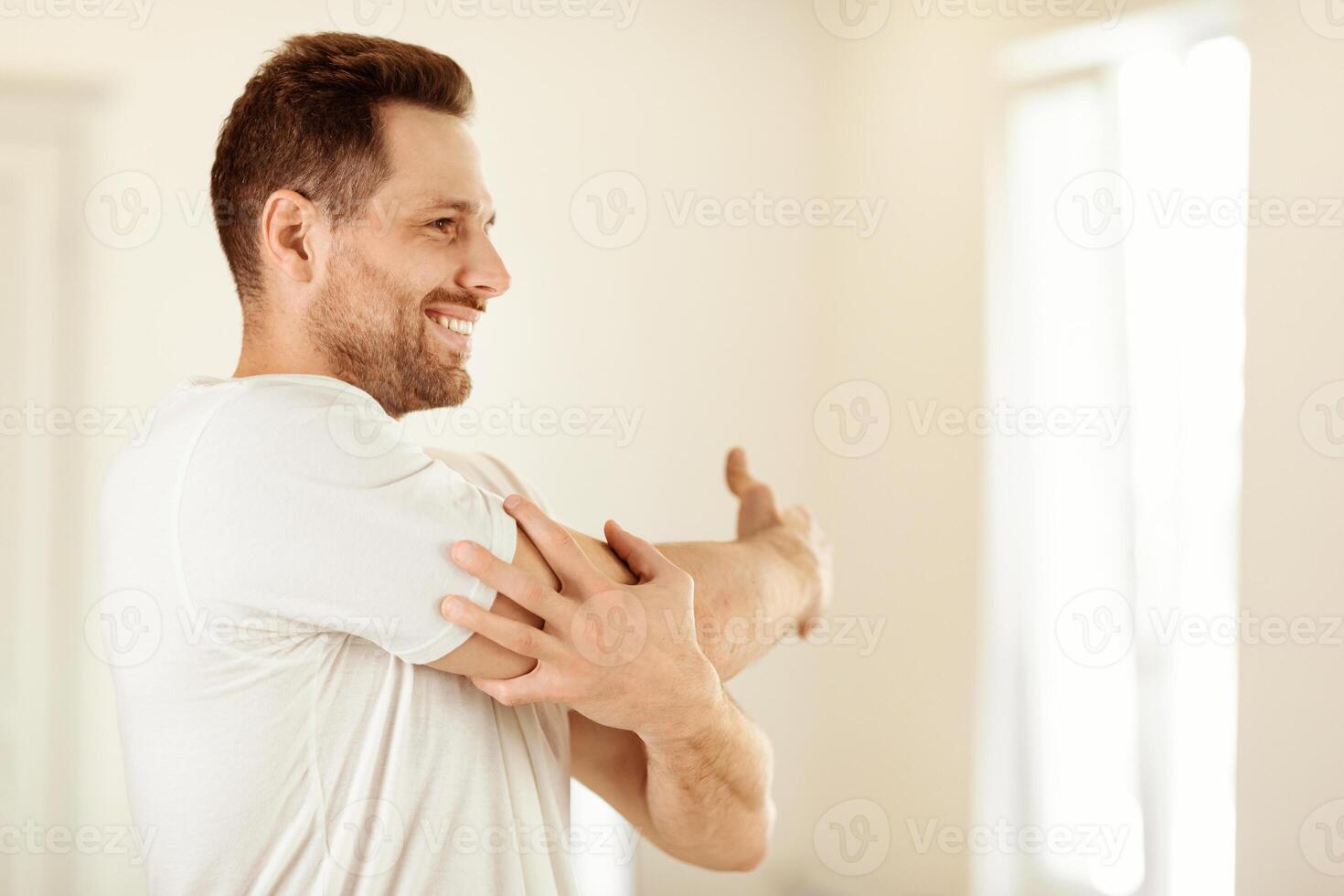 Cheerful Guy Exercising Stretching Arms Training Standing At Home. Morning Physical Exercises, Male Fitness, Daily Workout Routine Concept. Copy Space For Text photo