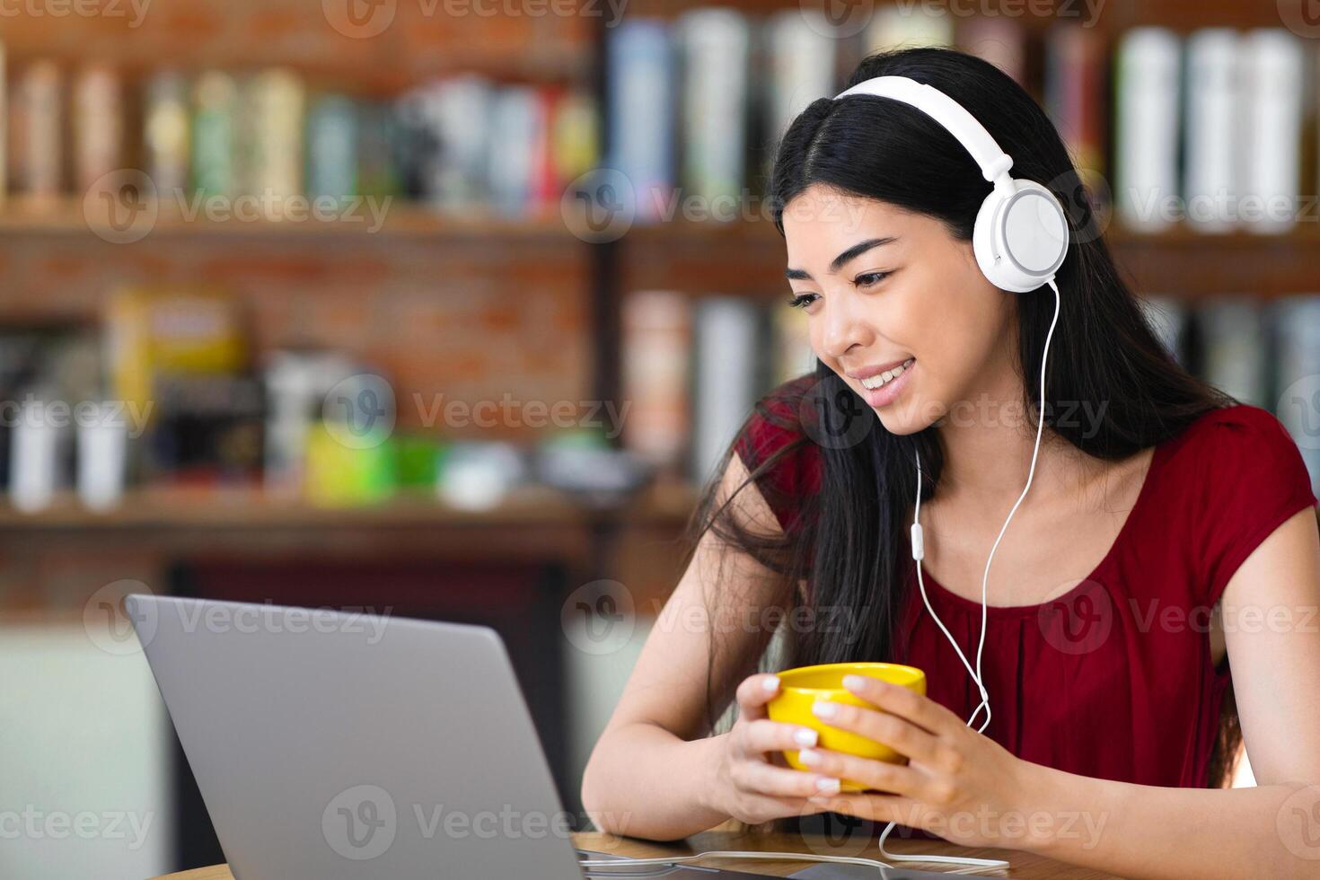 Smiling Asian Woman Watching Webinar With Laptop And Headphones In Cafe And Drinking Coffee, Having Conference Or Online Lesson, Looking AT Computer Screen And Smiling, Enjoying E-Learning photo