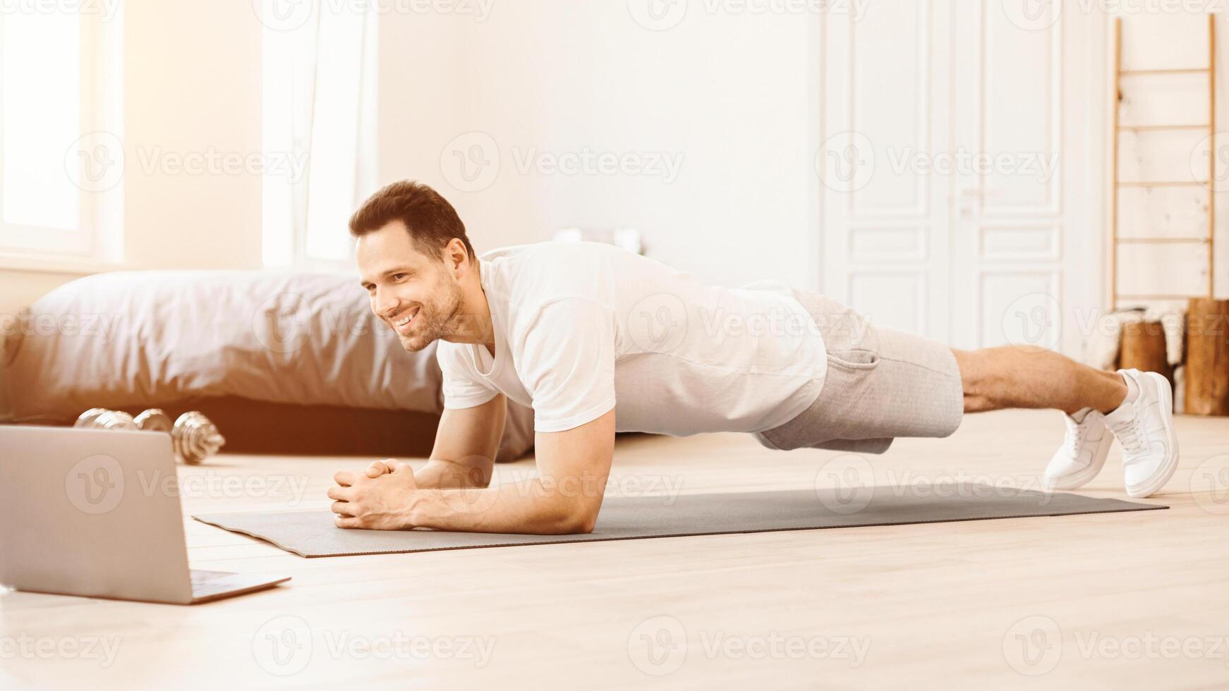 Online Workout. Happy Guy Doing Plank Exercise At Laptop Computer Training On Floor At Home. Panorama photo