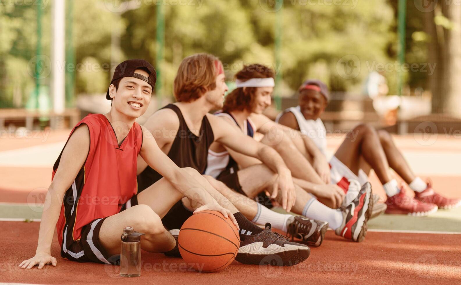 Smiling Asian basketball player with his diverse team sitting on outdoor game court, copy space. Panorama photo