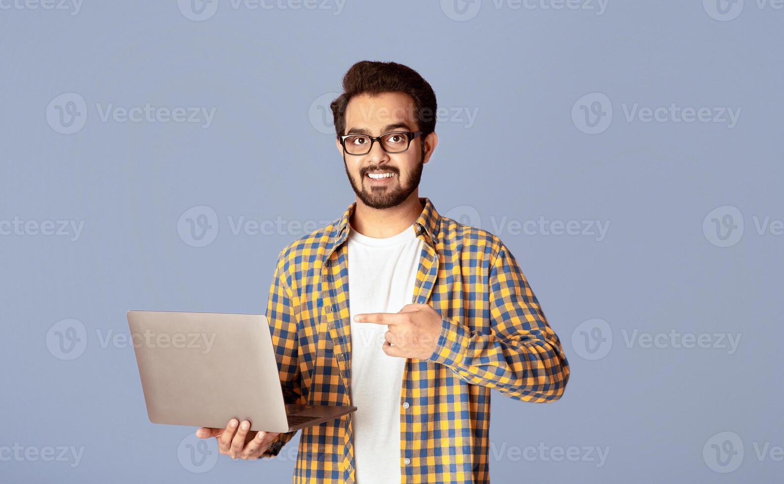 Online work, education, communication concept. Happy Indian guy pointing at laptop over lilac background photo