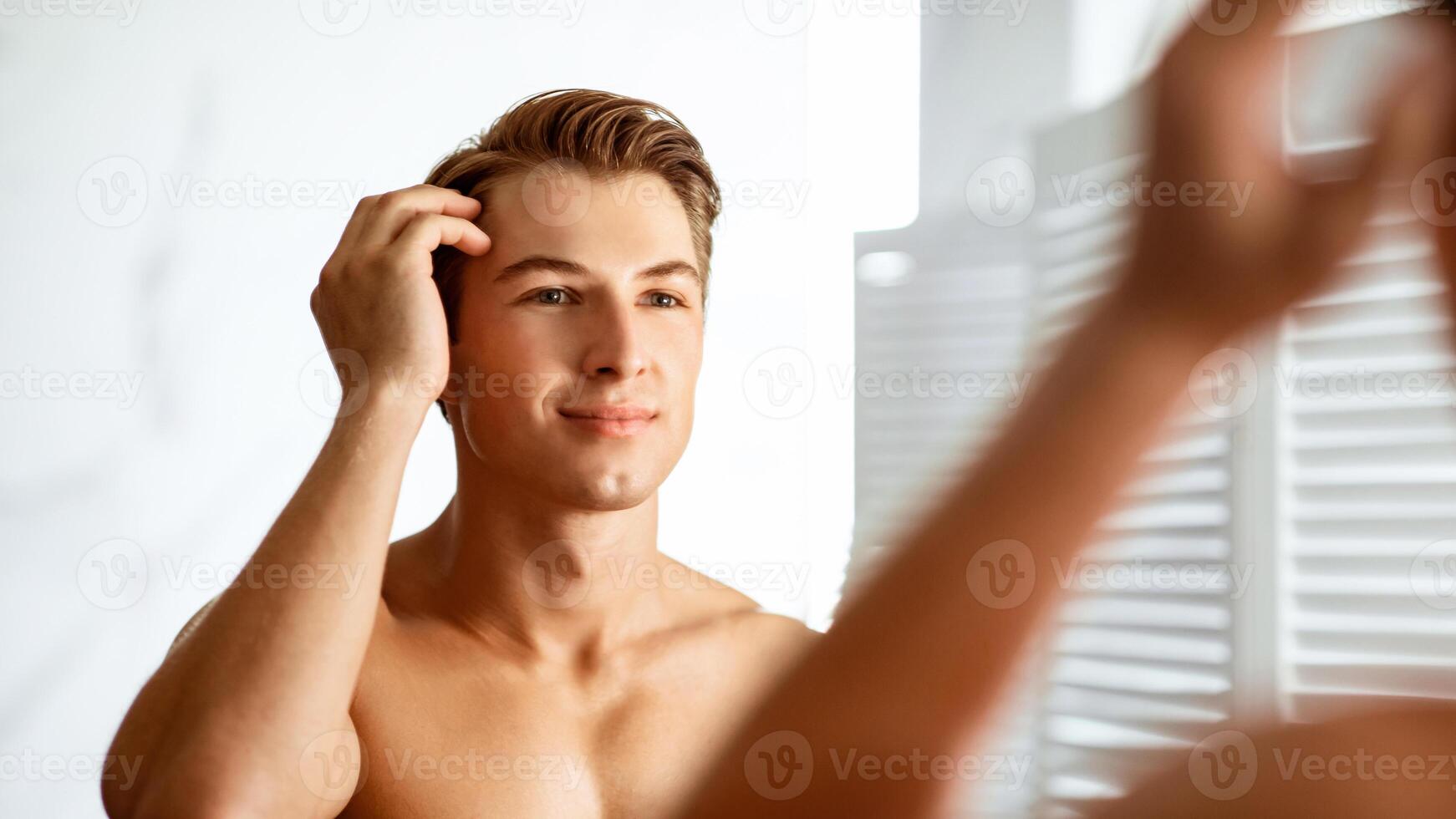 Confident Person Concept. Closeup portrait of charming man brushing his hair with hand, gazing at himself in the mirror photo