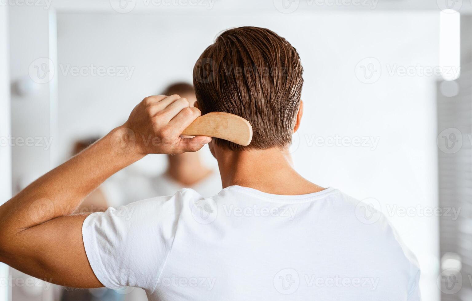 Male Haircare. Back view of unrecognizable young guy brushing his glossy hair with wooden comb, looking in the mirror at home or bathroom. Banner, copy space. Men's Beauty Routine Concept. photo