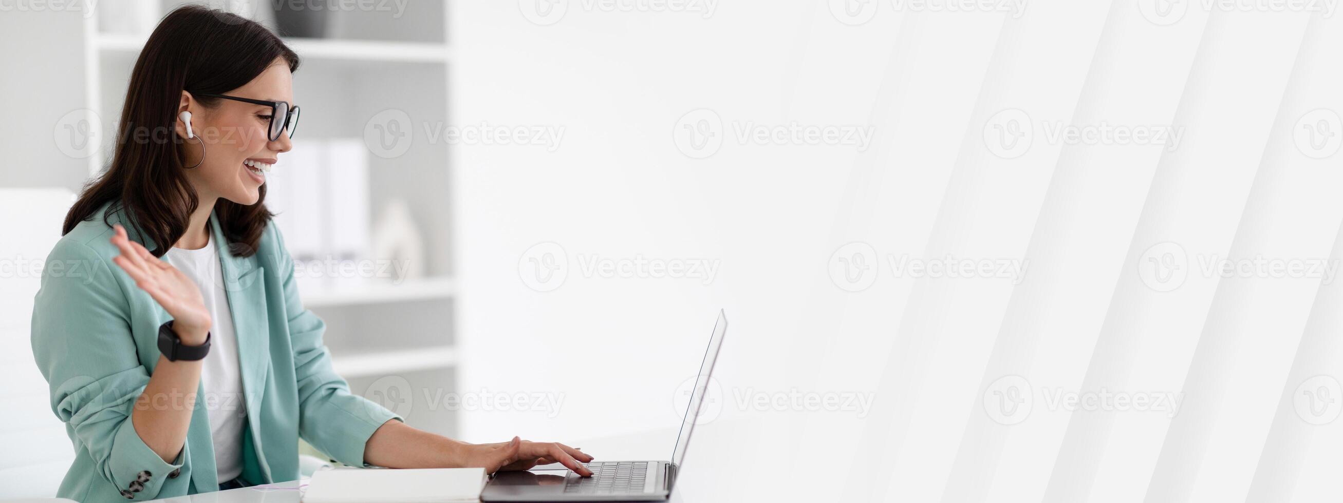 Smiling millennial caucasian lady in suit, glasses and wireless headphones waves hand at laptop at workplace in white office interior. Business with device, remote manager work, call, webinar photo