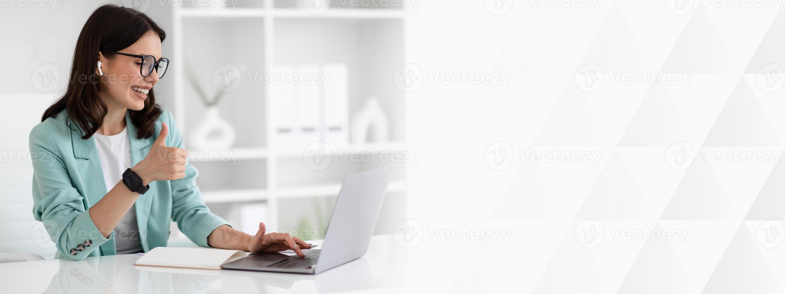Glad young caucasian businesswoman in suit at table typing on computer, show thumb up in office interior. Meeting remotely, approve business, work and technology, communication photo