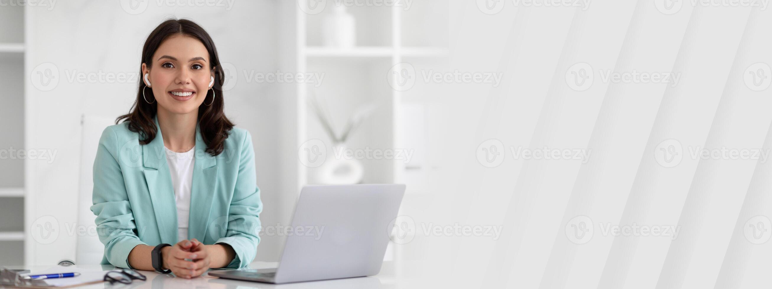 Glad young caucasian businesswoman in suit and wireless headphones at table with laptop in modern office interior. Meeting remotely, call, business, work with gadget alone photo