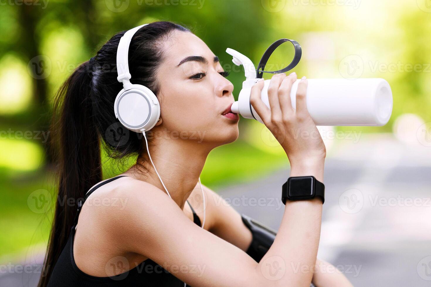 Thirsty After Training. Young asian girl drinking water from bottle, resting after jogging at green park, sitting on asphalt path, wearing headphones photo