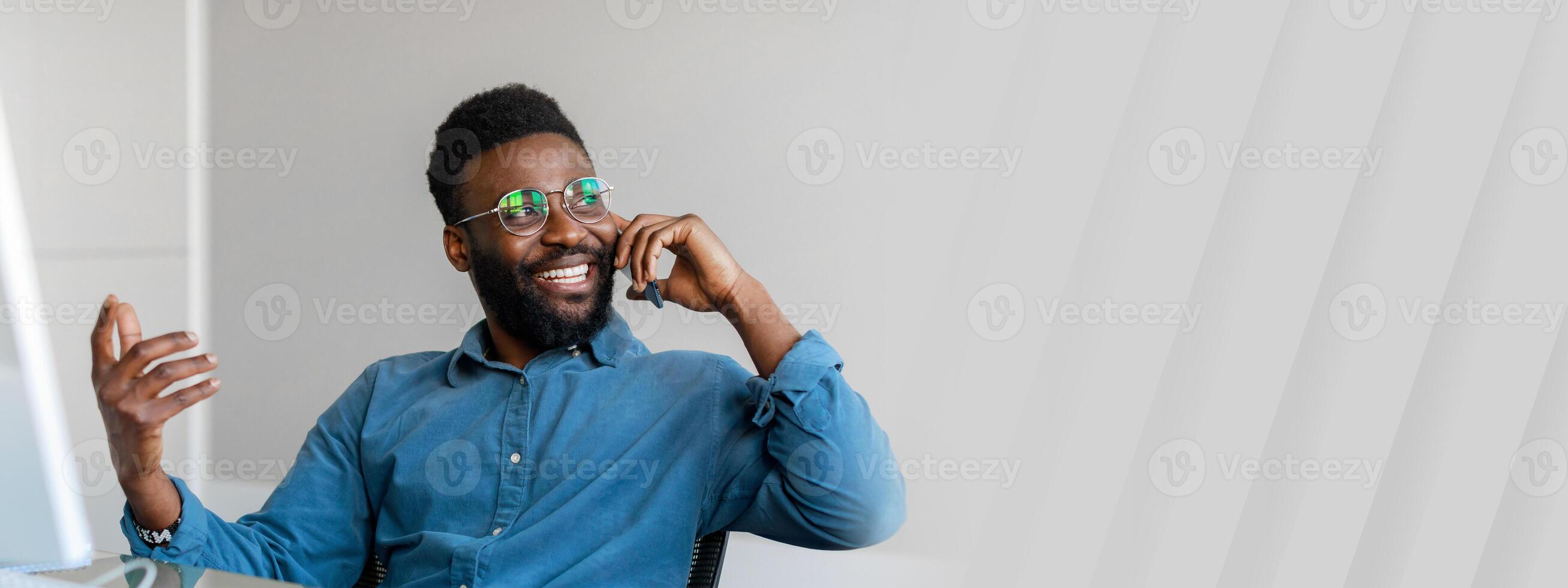 A young African American entrepreneur smiles while talking on the phone in a modern office. He demonstrates confidence and professionalism in a successful work environment, copy space photo