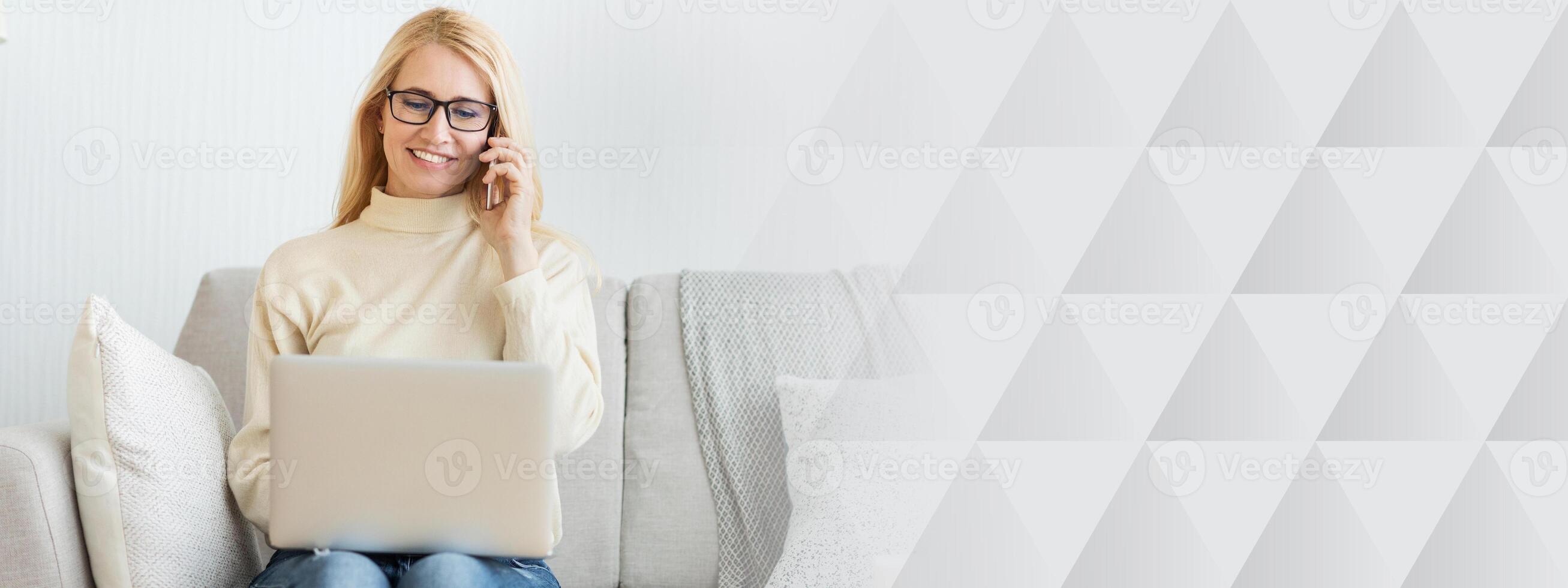 A woman sits comfortably on a sofa, talking on her phone while typing on her laptop. She appears relaxed in a cozy living room, with soft cushions and a warm ambiance, copy space photo
