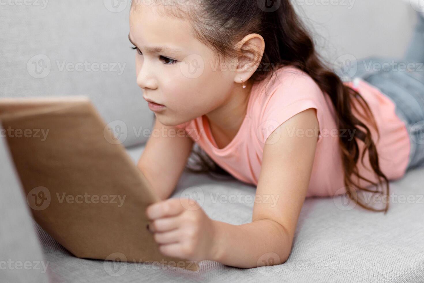 Little Asian Girl Reading Book Lying On Couch At Home. Kid's Weekend Leisure. Selective Focus photo