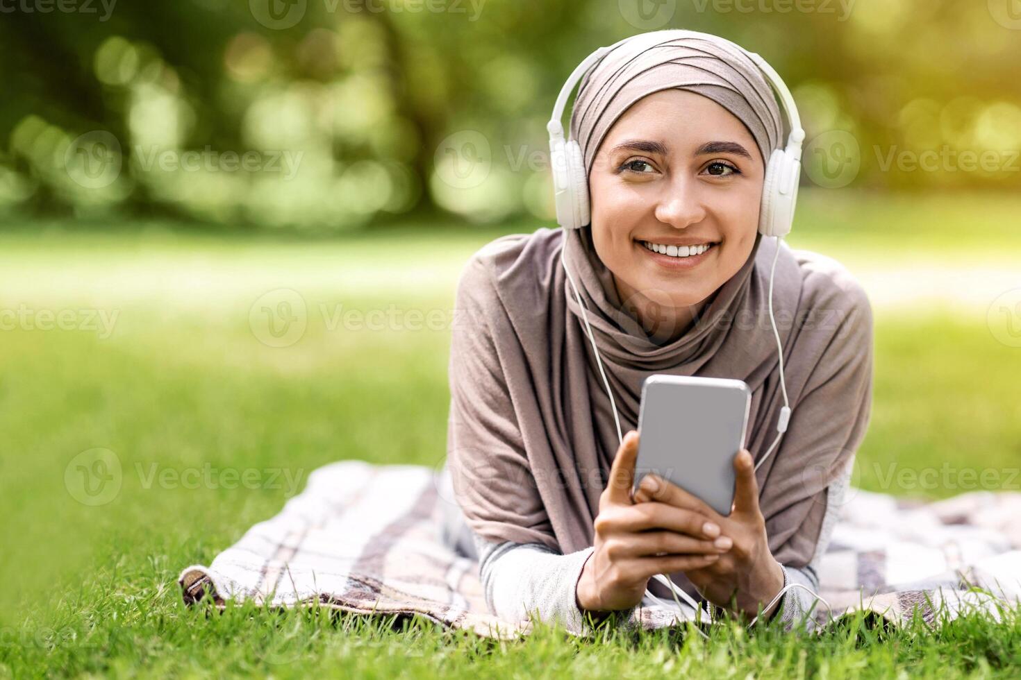 Positive muslim girl lying on blanket at park, using phone, watching , free space photo