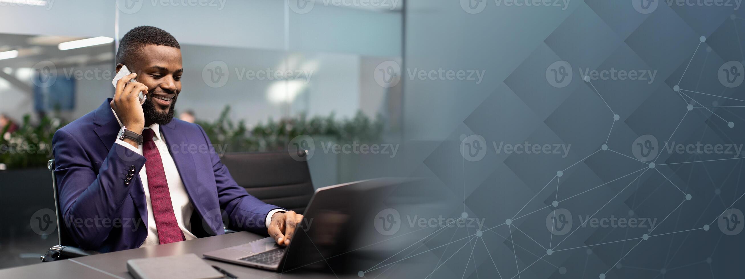 Successful african american young businessman in expensive blue suit working on laptop at office, having phone conversation with his personal assistant or business partner and smiling, copy space photo