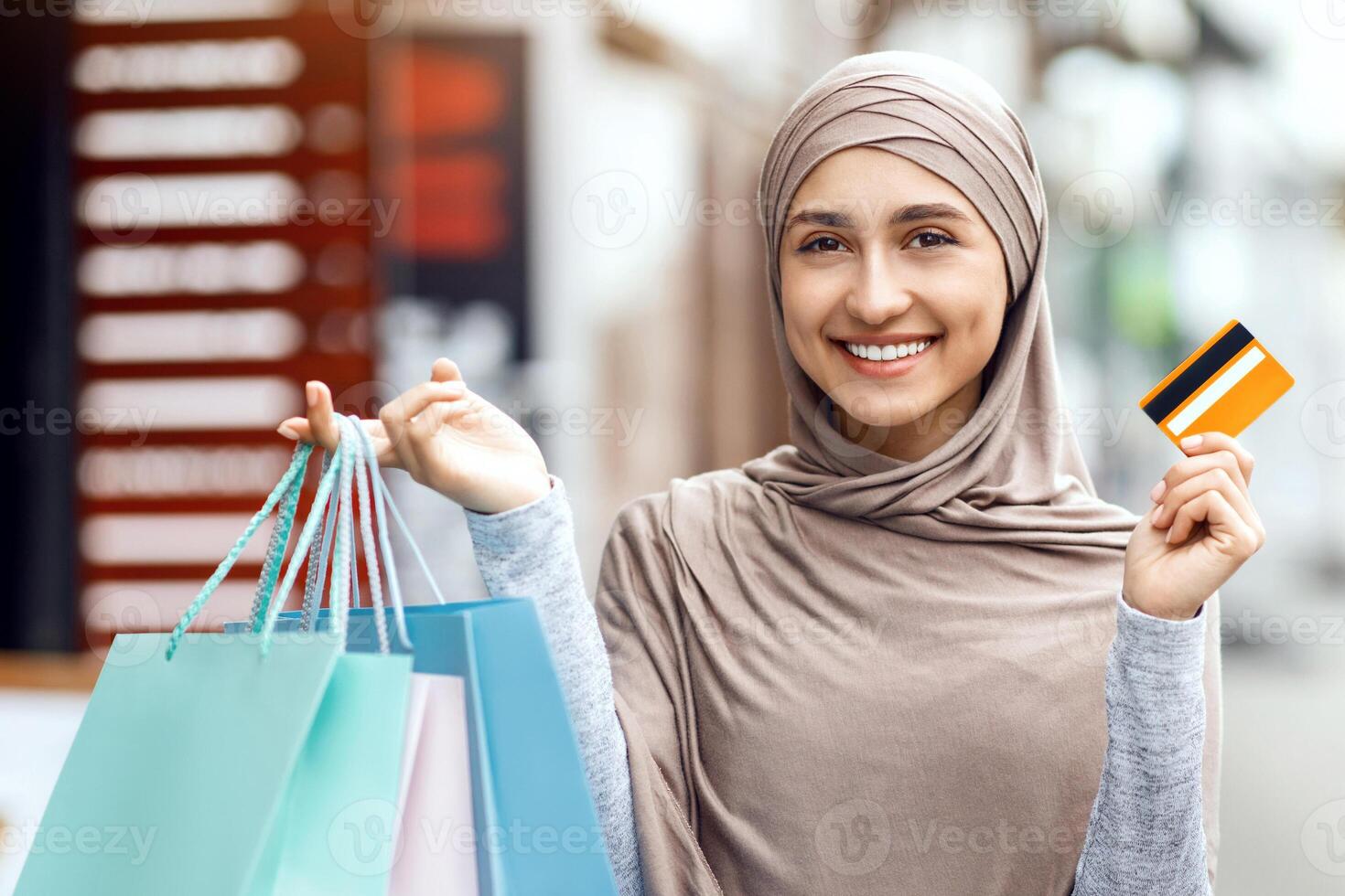 Shopaholic, shopping concept. Positive arab woman in hijab standing on street and showing shopping bags and credit card photo
