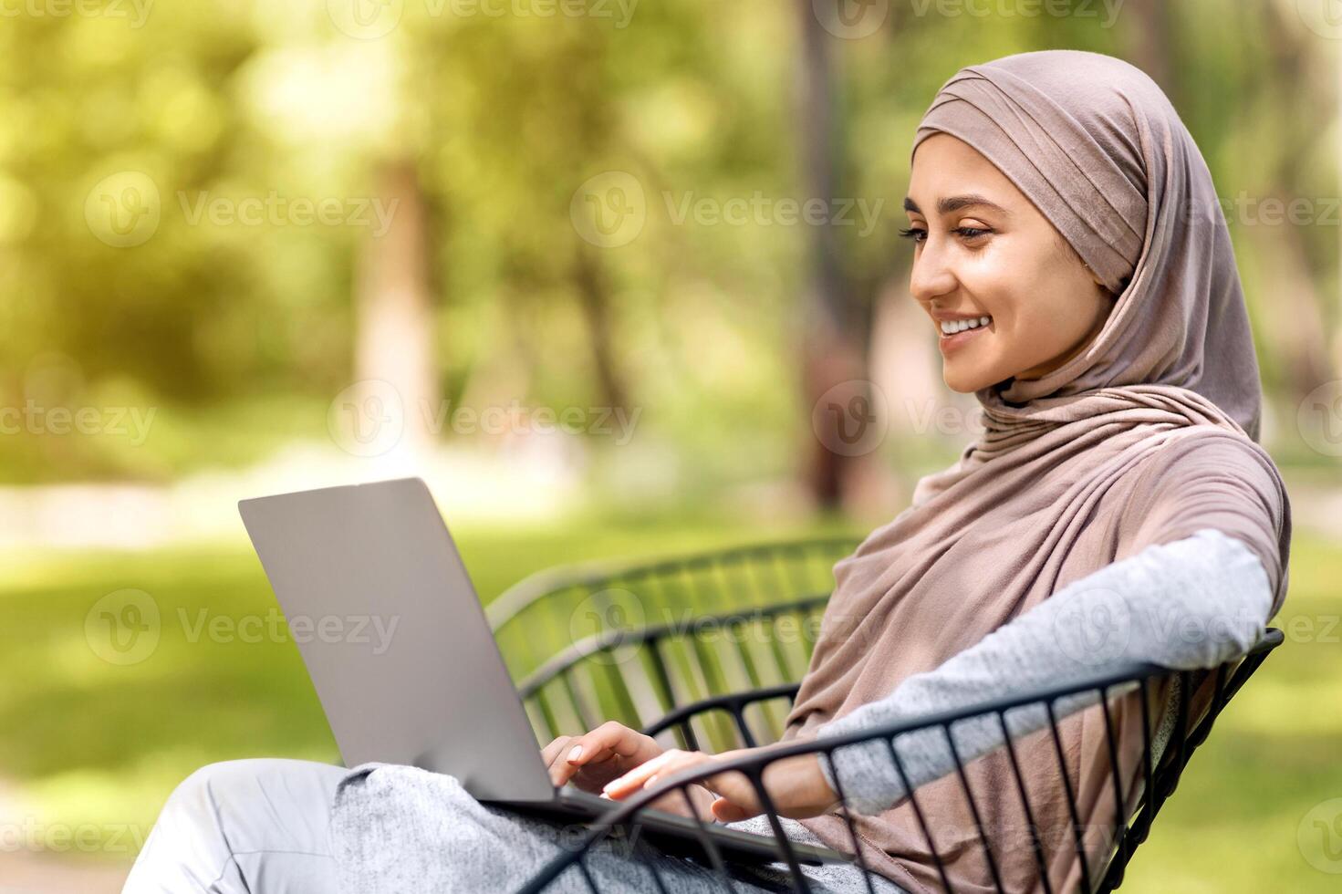 Young happy woman in hijab using laptop while sitting on bench at park, working outdoors, free space photo