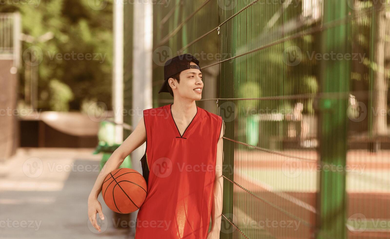 Millennial Asian basketball player with ball standing near fence at outdoor sports arena, panorama photo