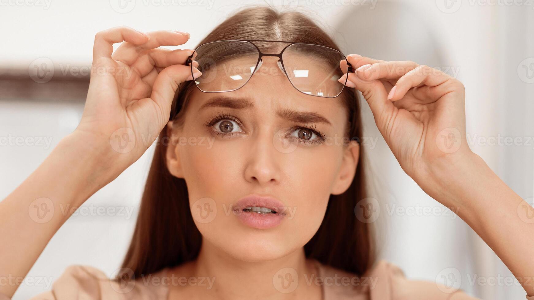 The woman appears puzzled as she holds her eyeglasses close to her face. Her expression reflects confusion, possibly about something she is viewing. The indoor setting is softly lit. photo