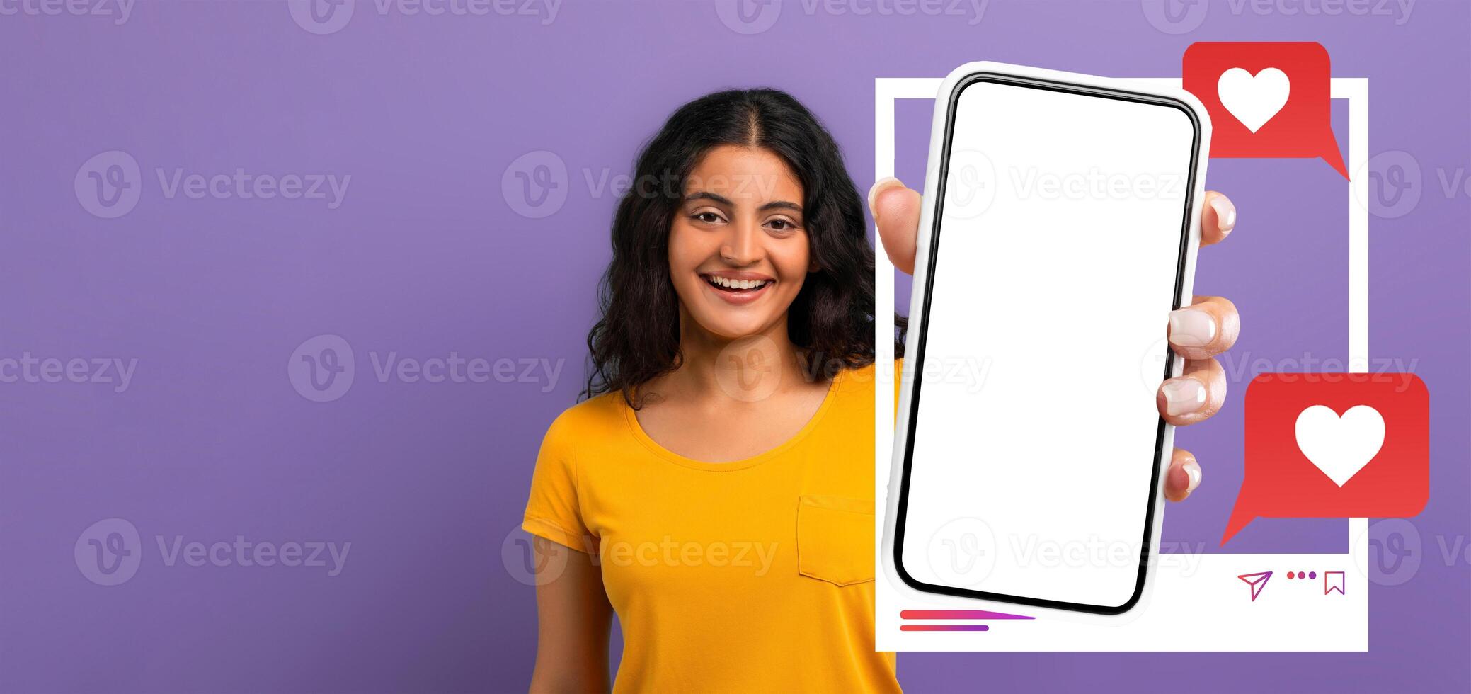 A young influencer smiles while holding a smartphone against a plain purple background. The phone displays a blank screen for social media applications, inviting interaction online. photo