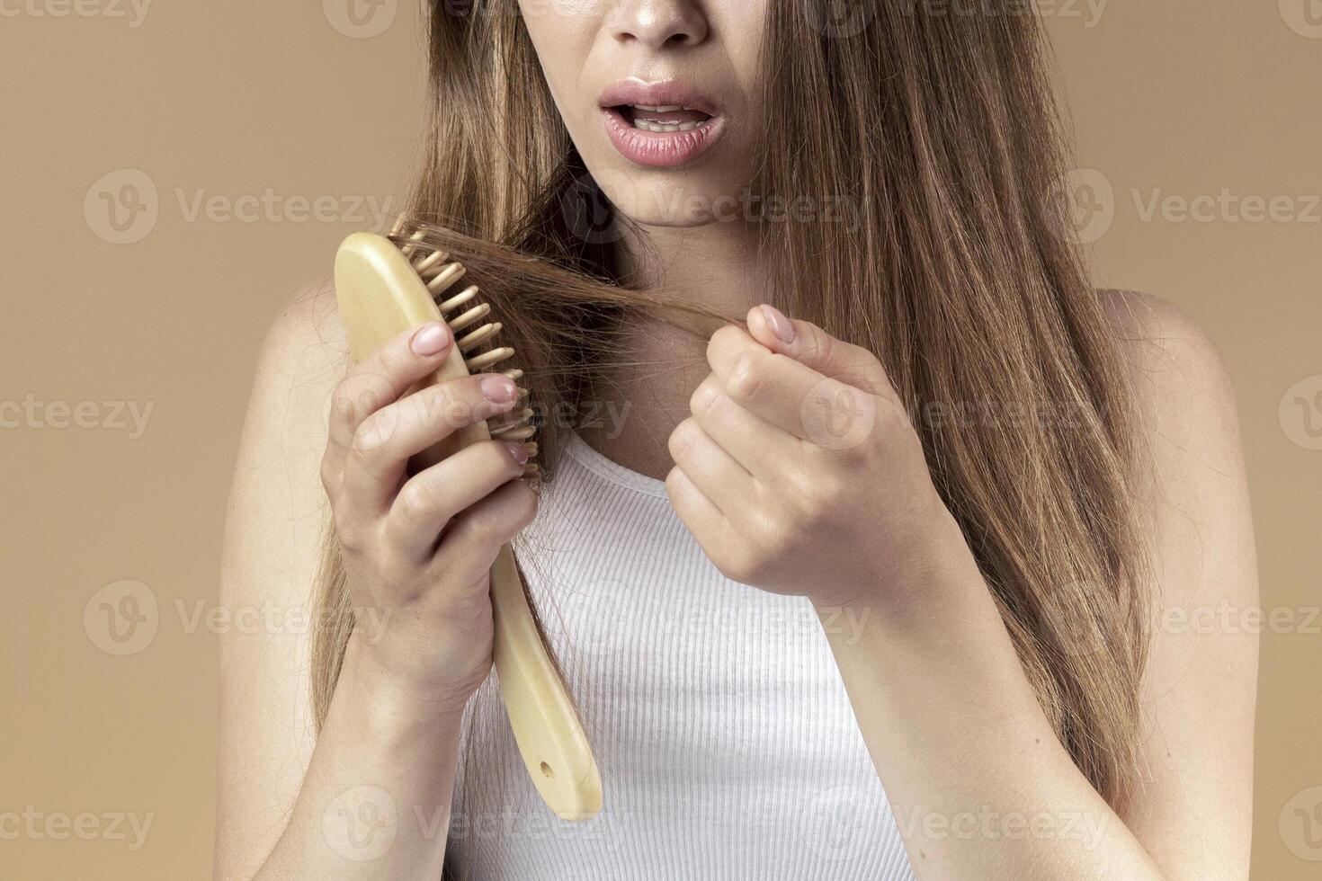 Frustrated young girl with wooden brush and disheveled hair on beige studio background photo