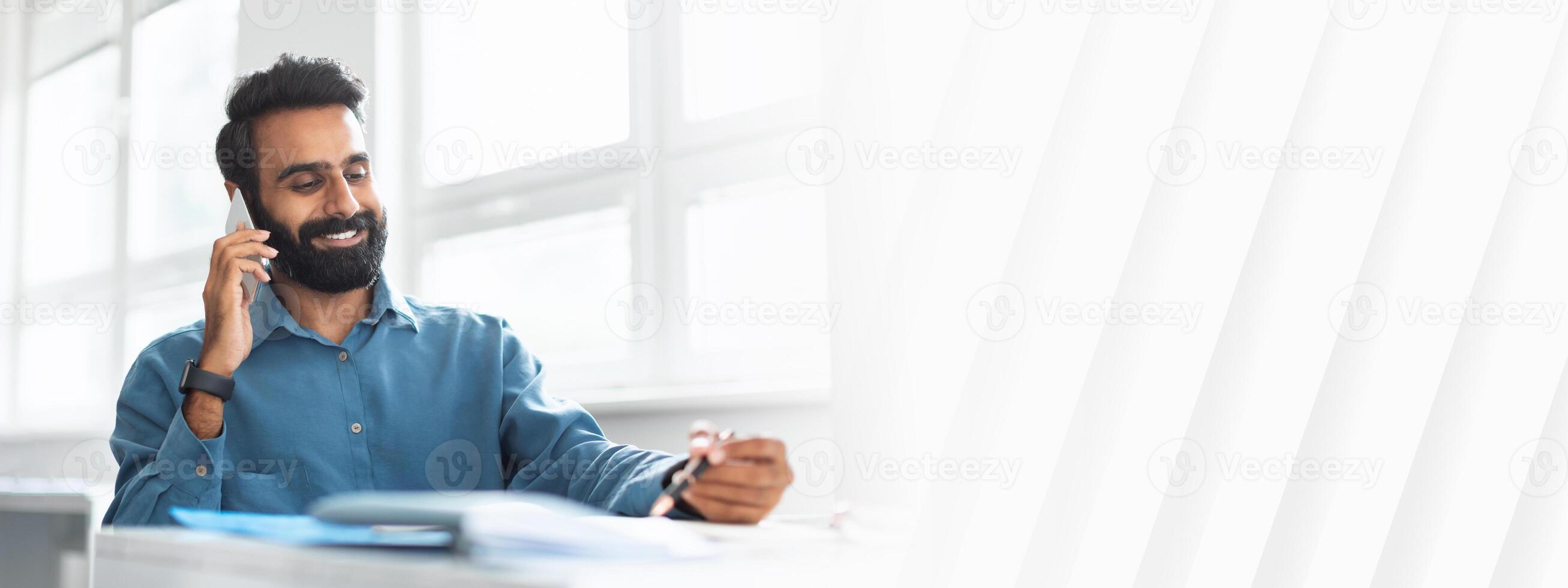 Smiling indian businessman talking on cellphone and noting down information, man entrepreneur speaking on mobile phone and taking notes, sitting at desk in office photo