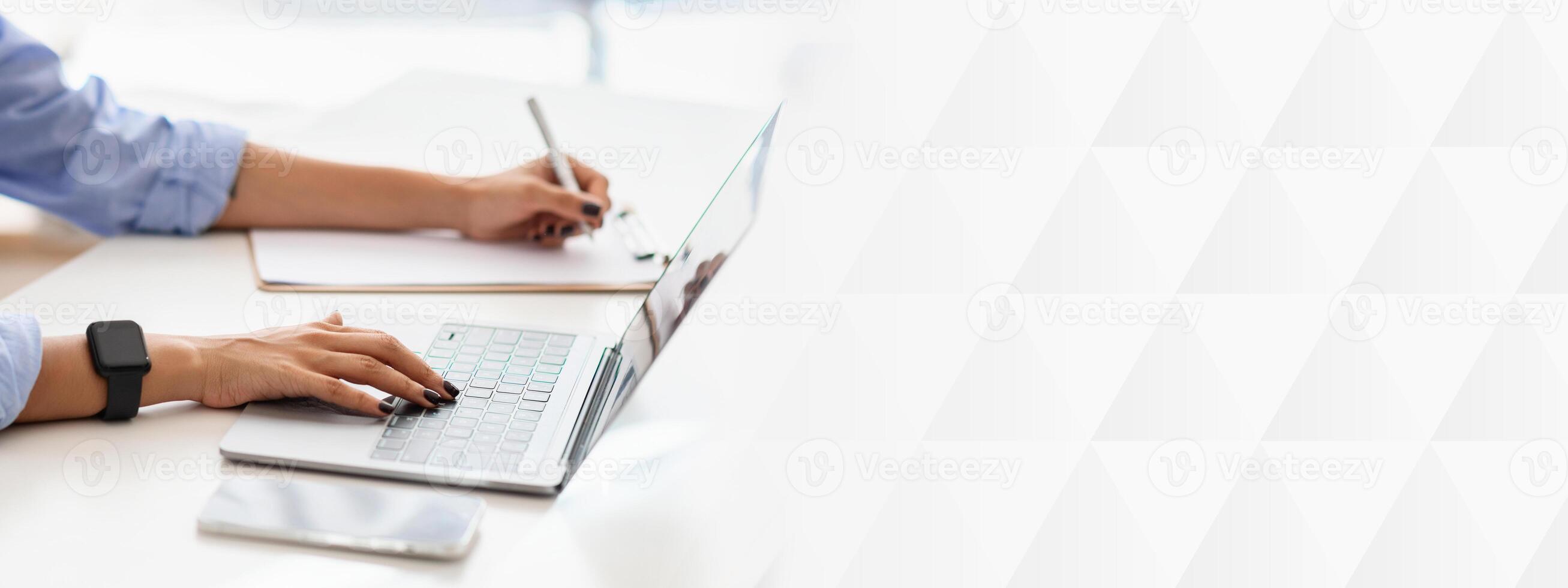 Professional woman in blue shirt multitasking, typing on a laptop while writing notes on a pad, wearing a smartwatch, focused on productivity and planning at white desk, cropped photo