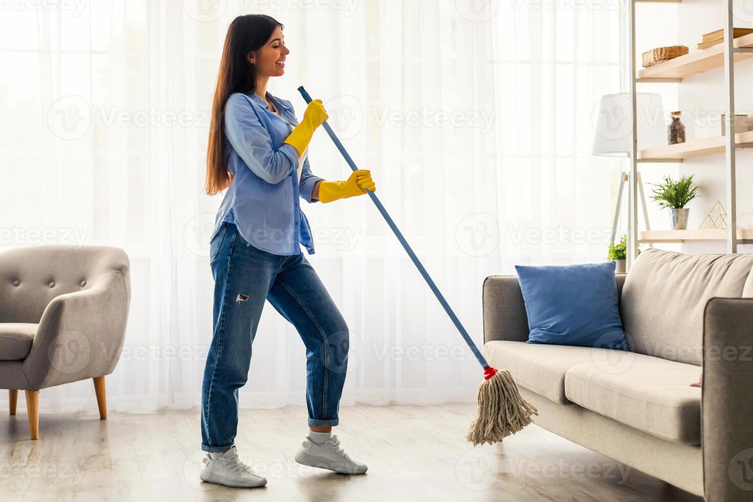 A young woman with long dark hair is happily cleaning her home. She is wearing yellow gloves, using a mop to clean the floor next to a couch in a bright living room. photo