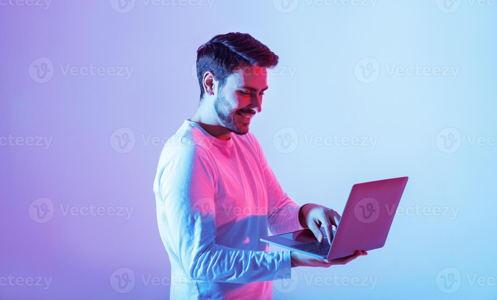A young man stands with a laptop, engaged in his work. The background is illuminated with vibrant pink and blue hues, creating a dynamic and modern atmosphere. photo