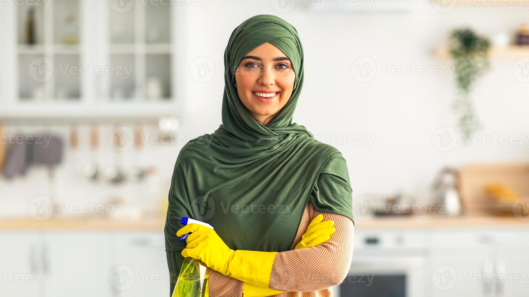 A young Muslim woman dressed in a green headscarf and yellow gloves is happily cleaning her kitchen. She holds a spray bottle and enjoys her time tidying up her space full of natural light. photo