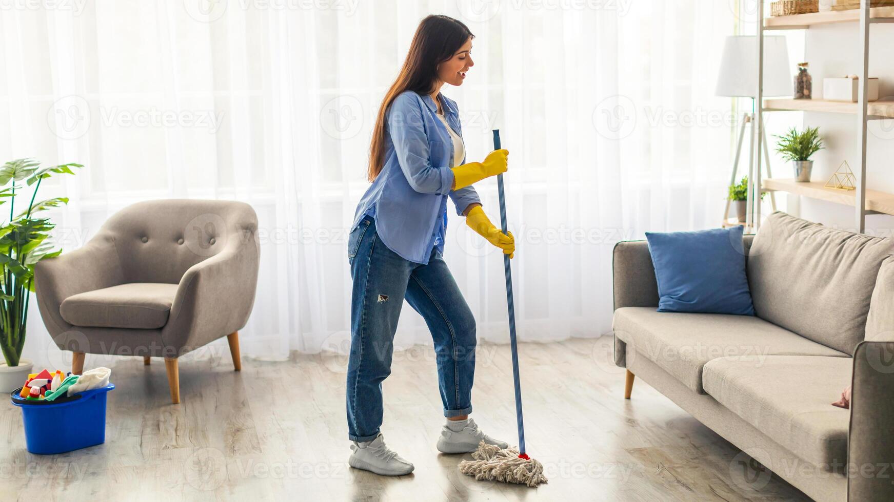 A young woman is mopping the floor in her cozy living room. She wears rubber gloves and casual clothes while enjoying a sunny atmosphere. The space is bright and inviting. photo