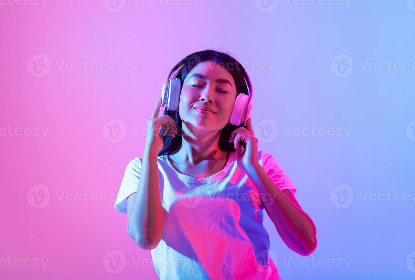 Asian woman is immersed in music while wearing headphones, with a serene expression on her face. The colorful background features vibrant pink and blue hues, enhancing her joyful moment. photo
