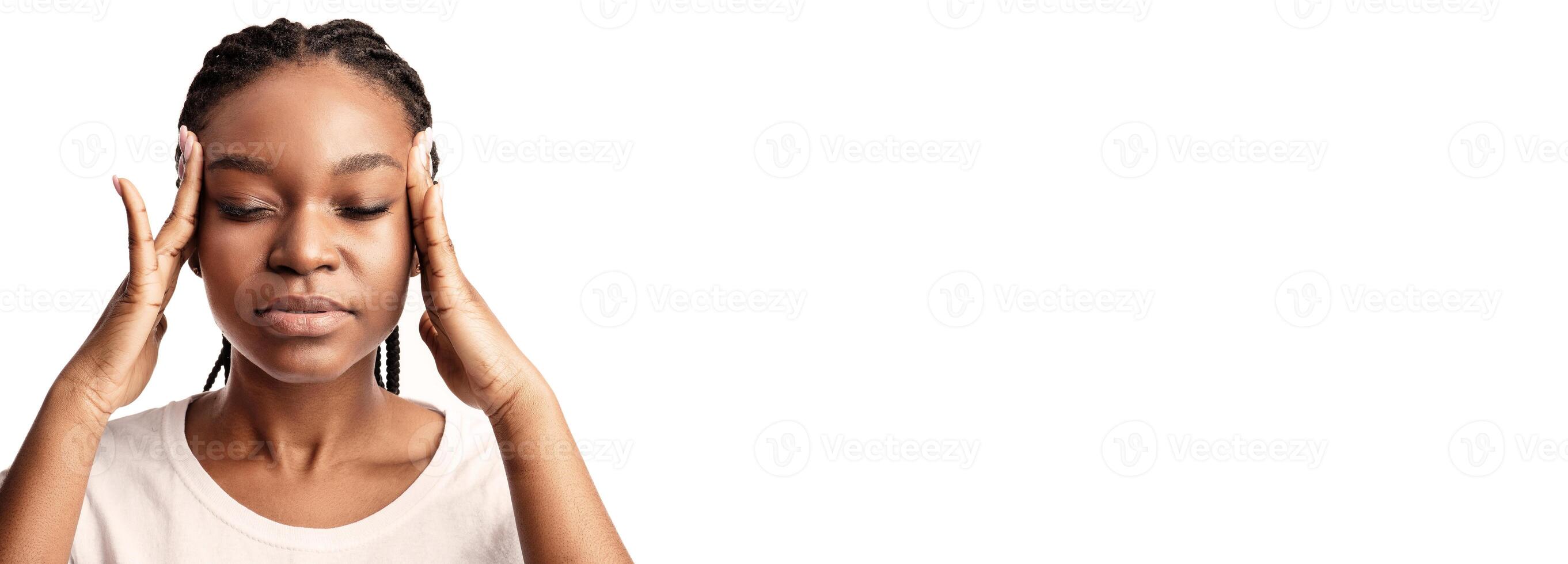 A young woman with braided hair is visibly stressed, holding her temples with both hands. The bright setting emphasizes her contemplative expression as she copes with overwhelming feelings. photo