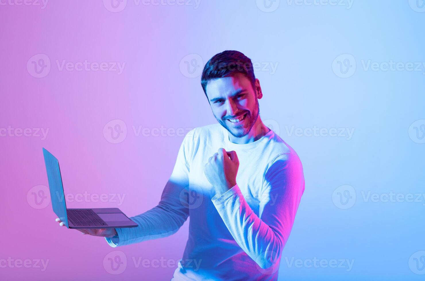 A young man in a light sweater expresses joy and triumph while using a laptop. The colorful background features shades of blue and pink, enhancing the celebratory atmosphere. photo