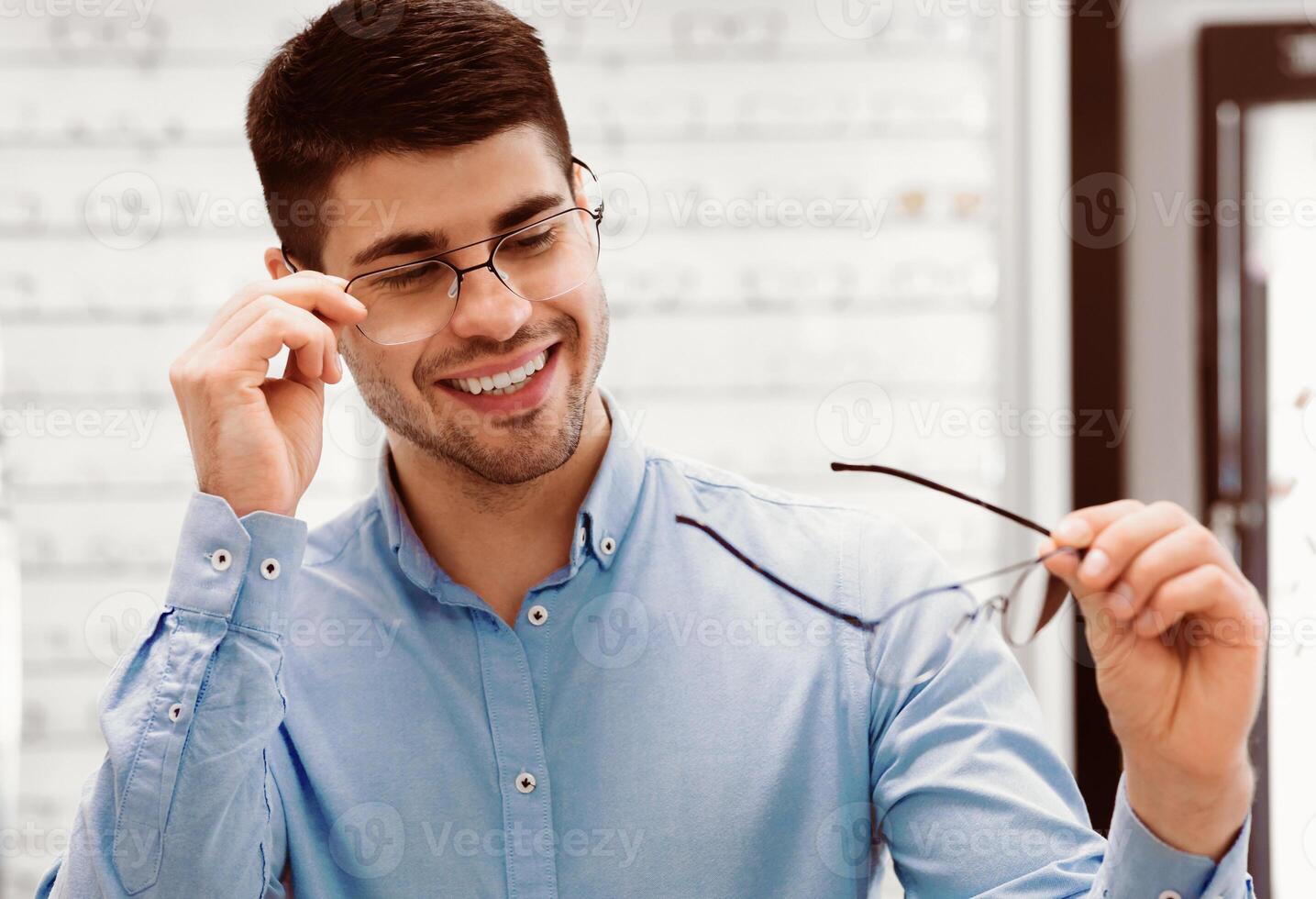 A man enjoys selecting new glasses while smiling in an eyewear store. He holds a pair of frames and examines them closely, showing a relaxed and cheerful demeanor in a bright setting. photo