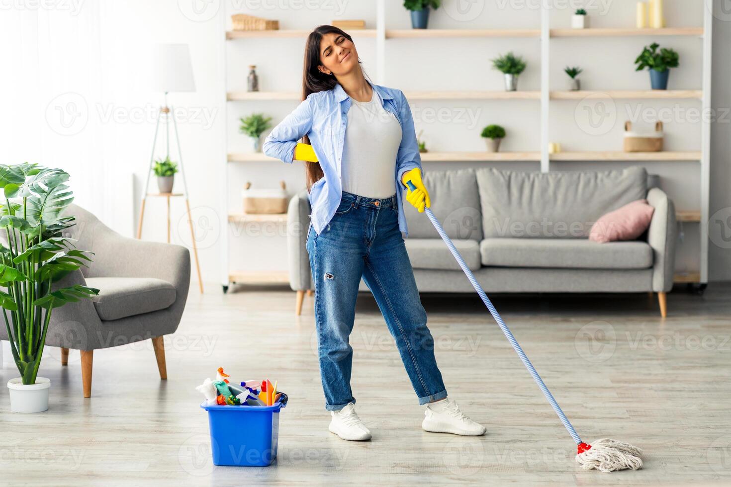 A young woman is cleaning her home, holding a mop with a tired expression. She pauses in a bright living room featuring plants, a sofa, and cleaning supplies in a bucket nearby. photo