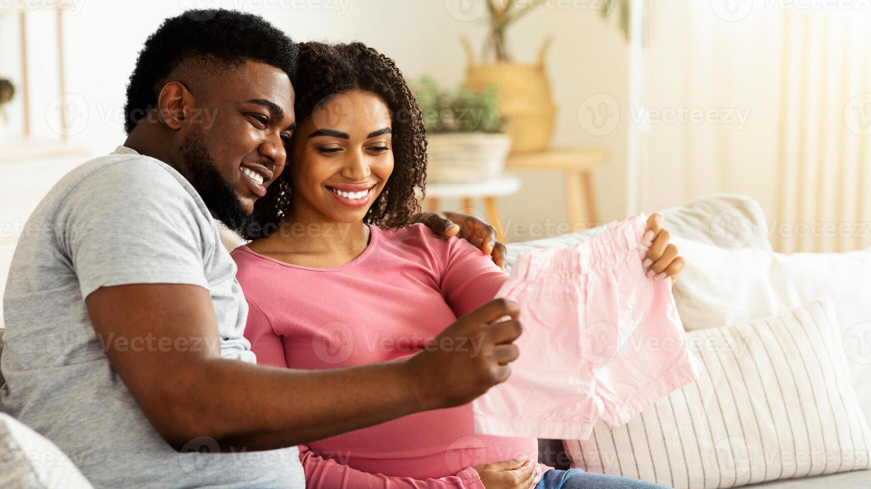 Smiling black expecting couple holding pink baby clothes, enjoying future parenthood, home interior, free space photo