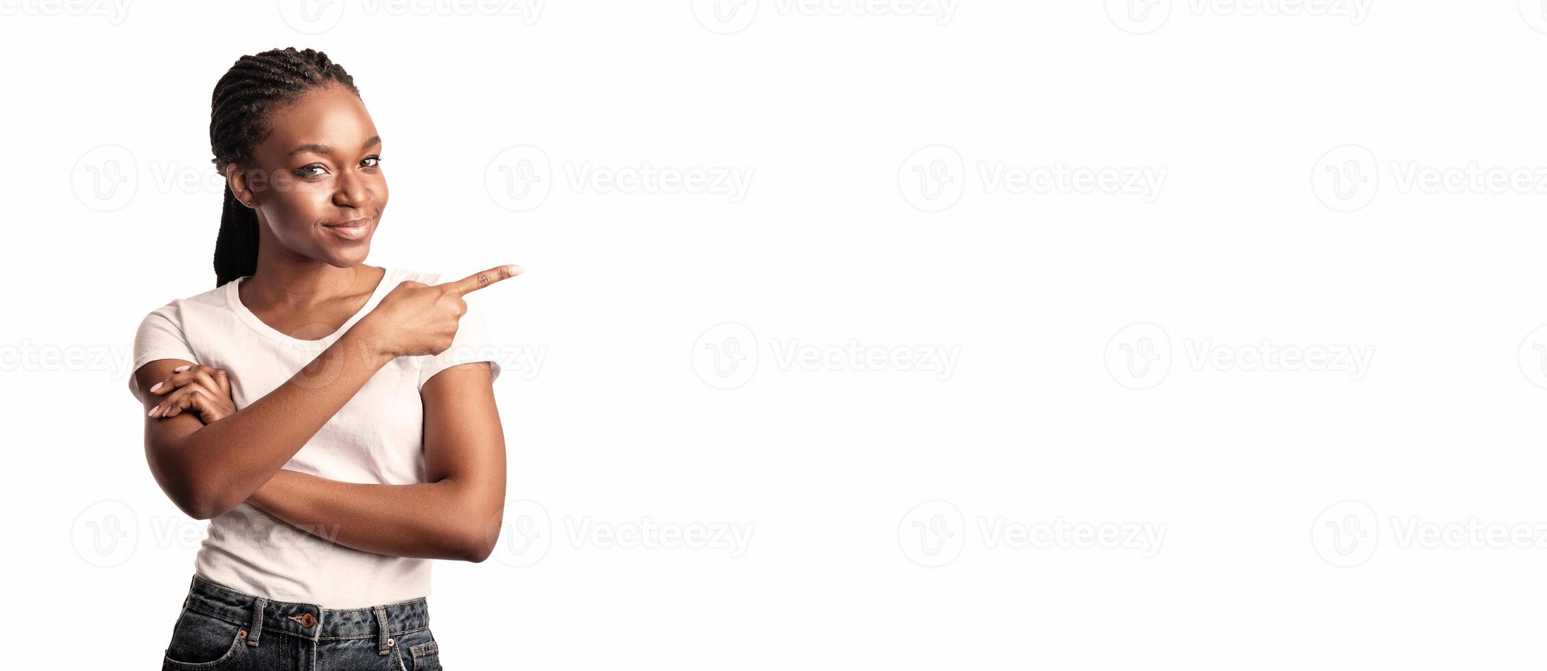 A young lady stands confidently with her arms crossed, smiling while pointing to the right. She is dressed in a simple t-shirt and denim shorts, suggesting a relaxed atmosphere. photo
