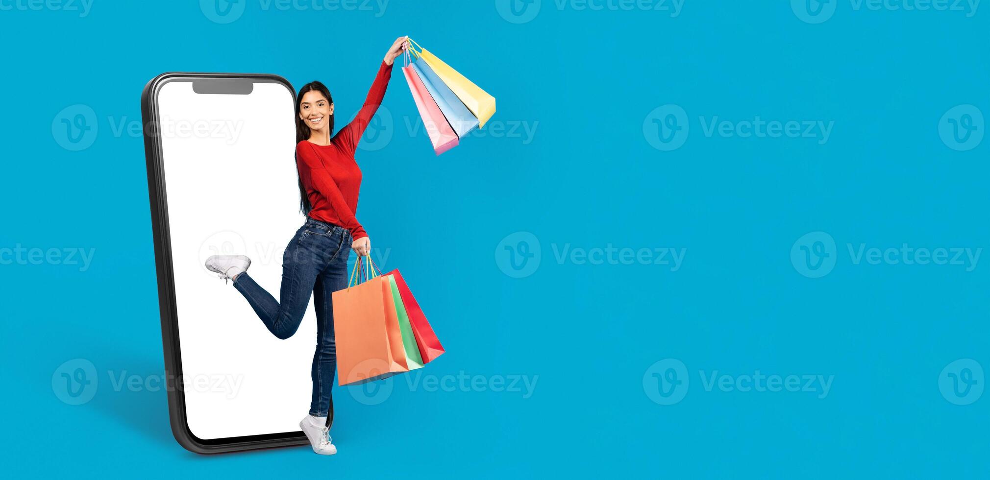 A cheerful customer holds shopping bags while stepping joyfully from a large smartphone screen. She embodies the excitement of online shopping and modern commerce in a vibrant setting. photo